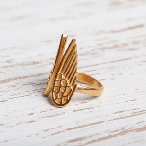May include: A gold-colored ring featuring a stylized wing design. The ring has a band and a detailed wing element with textured feather patterns. The wing is positioned vertically, creating a unique and artistic look. The ring is likely made of metal.