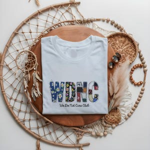 以下が含まれることがあります： 「WDNC We Do Not Care Club」の文字が入った水色のTシャツ。文字は花柄とパッチワーク模様でデザインされています。木製の表面に折りたたまれ、装飾品に囲まれています。