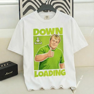 Puede incluir: Camiseta blanca con un diseño gráfico verde de un hombre haciendo una señal de pulgar hacia arriba, con las palabras "DOWN LOADING". El hombre lleva una camiseta verde. La camiseta está colgada en una percha negra.