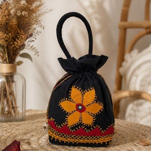 Könnte beinhalten: Schwarzer Beutel mit Kordelzug und einer großen, applizierten orange-gelben Blume. Die Tasche hat einen schwarzen Griff und ist mit roten und gelben Stickereien verziert. Die Tasche steht auf einer gewebten Matte.