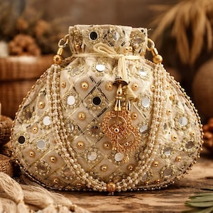 Puede incluir: Una bolsa adornada de color marfil con cierre de cordón. La bolsa está adornada con detalles dorados, pequeños espejos y cuentas de perlas. Una borla y un colgante decorativo dorados cuelgan de la parte delantera.