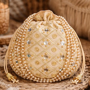 May include: A cream-coloured drawstring pouch with intricate gold embroidery and embellishments. The bag is adorned with pearls and sparkling accents, and features a decorative tassel on the drawstring.