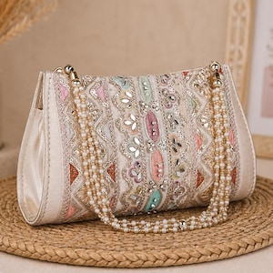 Op de afbeelding: Een rechthoekige clutch met ingewikkeld borduurwerk in roze, groene en gouden tinten, geaccentueerd met kleine spiegeltjes en lovertjes. De tas heeft een meerstrengs handvat gemaakt van kleine witte kunstparels en goudkleurige hardware.