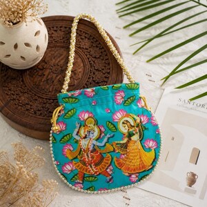May include: A turquoise drawstring bag with a beaded handle and a floral and figure design. The bag features a pattern of pink lotus flowers and green leaves. The figures are dressed in colorful traditional clothing.