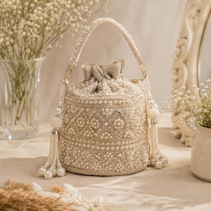 May include: An ivory-colored, beaded handbag with a drawstring closure and a braided handle. The bag is adorned with pearls and intricate beadwork, featuring dangling pearl tassels. The design is elegant and suitable for special occasions.