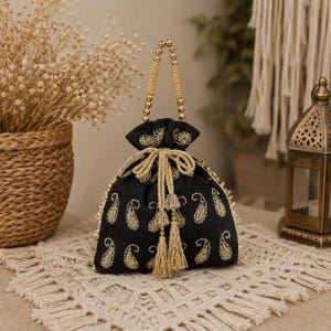 May include: Black drawstring bag with gold paisley embroidery and a beaded handle. The bag has gold tassels and trim, displayed on a cream fringed placemat. A decorative lantern and basket are in the background.