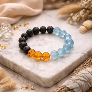 May include: A beaded bracelet featuring black lava rock beads, amber-colored beads, and light blue beads. The bracelet is displayed on a white marble surface, with decorative elements in the background.