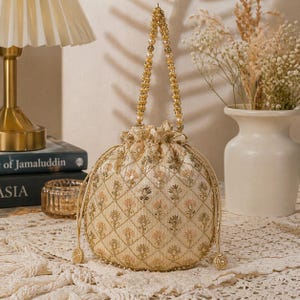 Puede incluir: Un bolso con cordón bordado de color crema con un asa dorada con cuentas. El bolso presenta un patrón de diamantes con detalles florales y lentejuelas doradas. El asa está adornada con pequeñas perlas. El bolso está sobre una superficie texturizada de color crema.