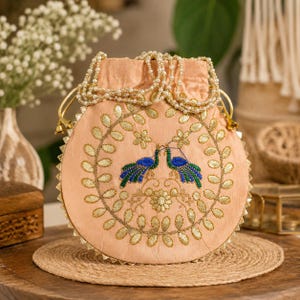 Puede incluir: Una bolsa redonda bordada de color melocotón con cierre de cordón. La bolsa presenta un diseño de hojas y flores doradas con dos pavos reales azules y verdes. El cordón está adornado con perlas y detalles dorados.