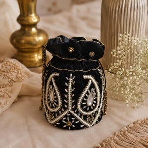 May include: Black velvet drawstring bag with detailed white and gold beaded paisley patterns. The bag has a beaded drawstring and is adorned with clear, teardrop-shaped crystals. A luxurious, decorative accessory.