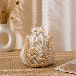 Può includere: Una borsa a coulisse bianca e dorata con perline, con interno color crema. La borsa è adornata da più fili di perline bianche e dorate, e nappe dorate pendono dai lati. La borsa è appoggiata su una superficie tessuta.