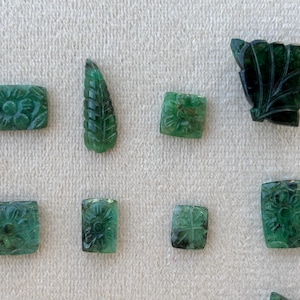 May include: A collection of carved emerald gemstones. The gemstones are in various shapes, including rectangular and leaf-shaped, and feature floral and leaf designs. The emeralds are a deep green colour and are displayed on a light-coloured surface.