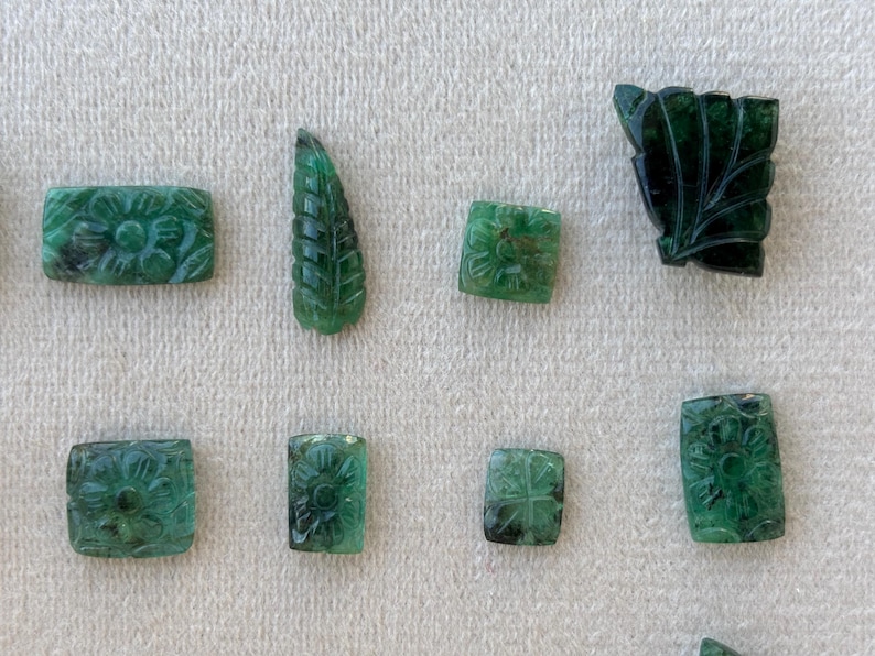 May include: A collection of carved emerald gemstones. The gemstones are in various shapes, including rectangular and leaf-shaped, and feature floral and leaf designs. The emeralds are a deep green colour and are displayed on a light-coloured surface.