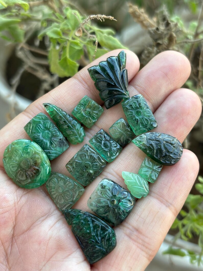 May include: A selection of carved emerald gemstones in diverse shapes, such as ovals, squares, and leaf designs. The gemstones are a rich green hue and display detailed floral and leaf carvings. The gemstones are held in a hand.