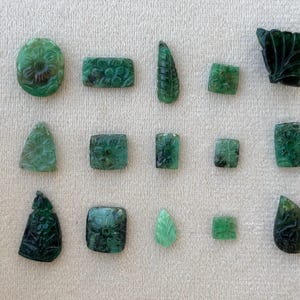 May include: A collection of carved emerald gemstones in diverse shapes, such as oval, rectangular, and square. The gemstones showcase floral and leaf patterns, with varying shades of green. The display is arranged on a white surface.