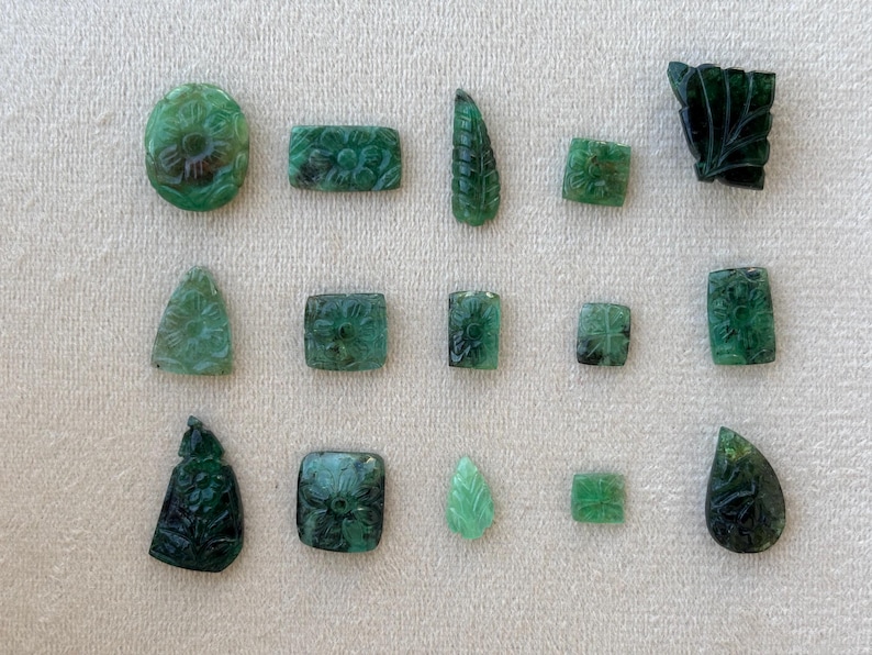 May include: A collection of carved emerald gemstones in diverse shapes, such as oval, rectangular, and square. The gemstones showcase floral and leaf patterns, with varying shades of green. The display is arranged on a white surface.