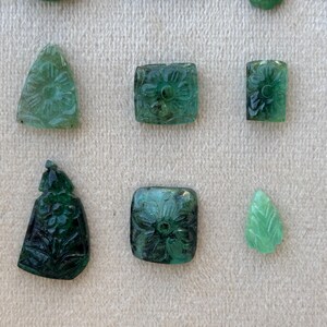 May include: A collection of carved emerald gemstones in diverse shapes, including triangles, squares, and leaf-like forms. The emeralds are a deep green colour and feature floral and geometric designs.