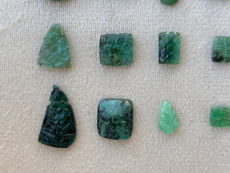 May include: A collection of carved emerald gemstones in diverse shapes, including triangles, squares, and leaf-like forms. The emeralds are a deep green colour and feature floral and geometric designs.