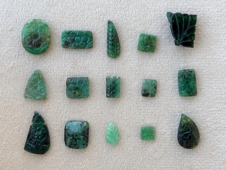 May include: A selection of carved emerald gemstones in diverse shapes, such as oval, rectangular, and leaf-like. Each gemstone displays detailed floral or leaf patterns, highlighting the natural green hue and intricate craftsmanship.