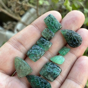 May include: A selection of carved emerald gemstones in various shapes, including squares, rectangles, and teardrops. The gemstones feature intricate floral and leaf designs. The emeralds are a deep green colour and are held in a hand.