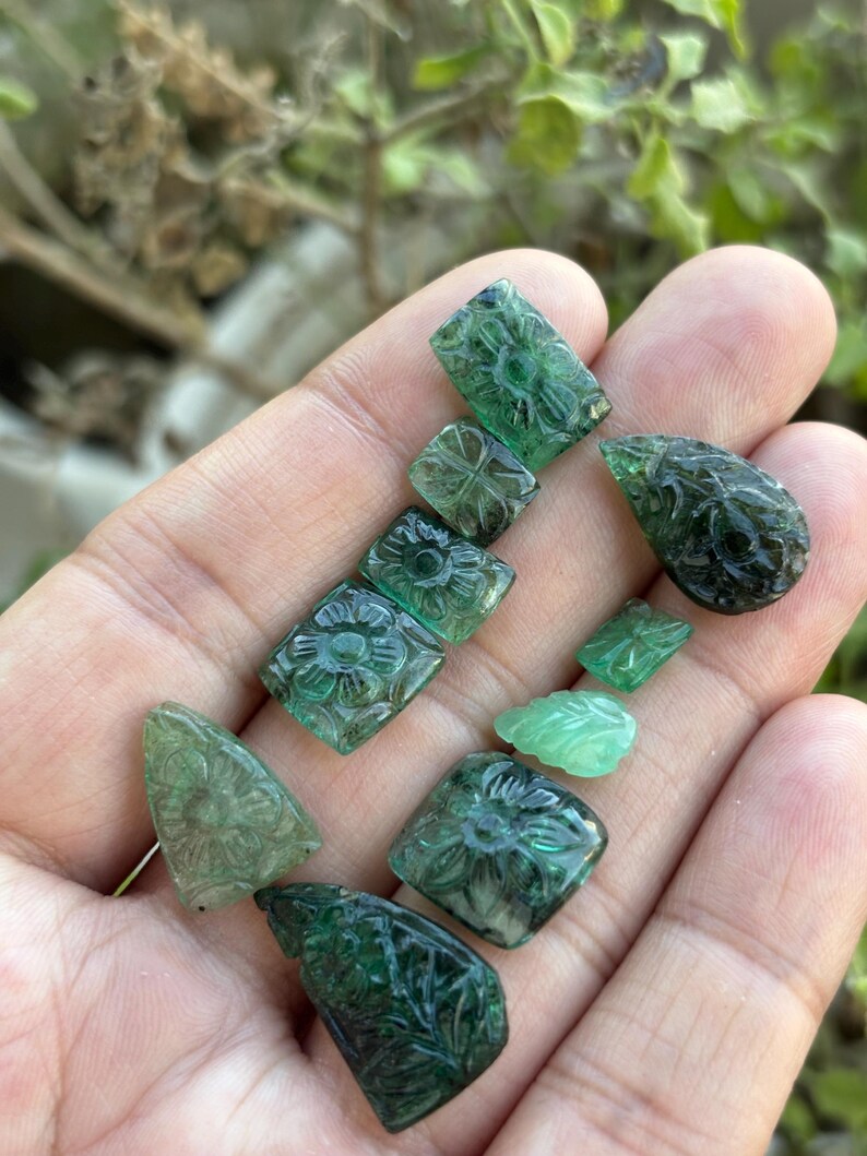 May include: A selection of carved emerald gemstones in various shapes, including squares, rectangles, and teardrops. The gemstones feature intricate floral and leaf designs. The emeralds are a deep green colour and are held in a hand.
