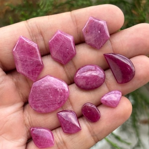 May include: A collection of ten loose, polished pink and magenta gemstones in various shapes, including hexagonal, oval, teardrop, and irregular forms. Each stone displays natural internal inclusions and a smooth, reflective surface texture.