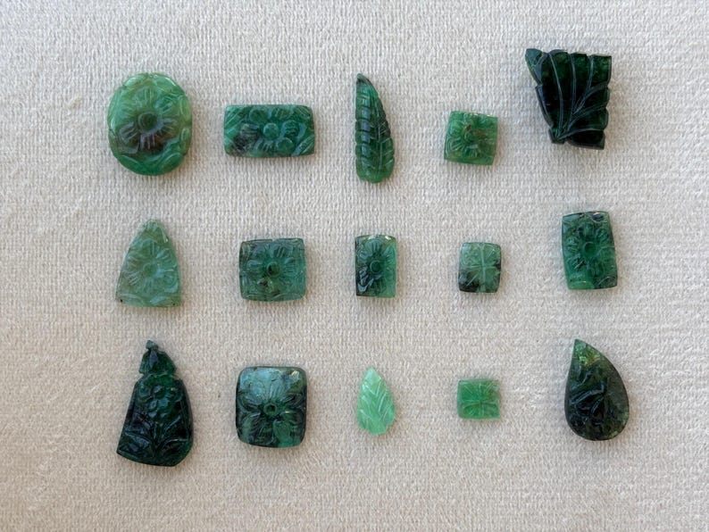 May include: A selection of carved emerald gemstones in diverse shapes, such as oval, rectangular, square, and teardrop. The gemstones display floral and leaf patterns, highlighting intricate details and a deep green hue.