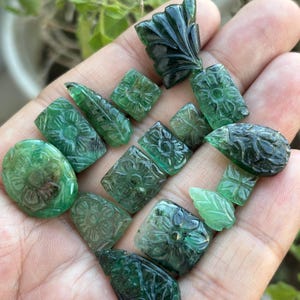 May include: A selection of carved emerald gemstones in diverse shapes, such as ovals, rectangles, and leaf forms. The gemstones are a deep green hue and display floral and leaf patterns. The gemstones are held in a hand.