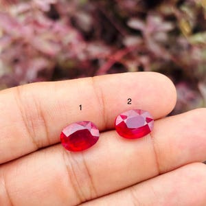 May include: Two oval-cut, vibrant red gemstones are displayed. The gemstone on the left is labelled with the number 1, and the gemstone on the right is labelled with the number 2. Both stones feature faceted surfaces that reflect light, showcasing a deep red colour.