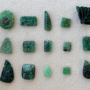 May include: A selection of carved emerald gemstones in diverse forms, such as oval, rectangular, and leaf-shaped. The gemstones display floral and leaf patterns, exhibiting intricate detail and a deep green hue.