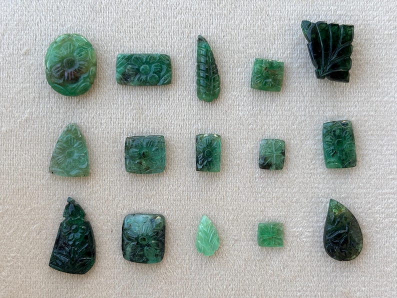 May include: A selection of carved emerald gemstones in diverse forms, such as oval, rectangular, and leaf-shaped. The gemstones display floral and leaf patterns, exhibiting intricate detail and a deep green hue.