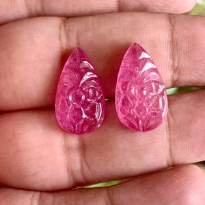 Puede incluir: Dos piedras preciosas rosas en forma de lágrima con un intrincado patrón floral tallado en la superficie. Estas piedras translúcidas exhiben un tono magenta vibrante y un acabado pulido, mostrando grabados detallados en un diseño simétrico.
