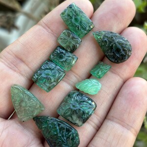 May include: A selection of carved emerald gemstones in various shapes, including squares, teardrops, and leaves. The gemstones are a deep green colour and feature intricate floral and leaf designs. The gemstones are held in a hand.