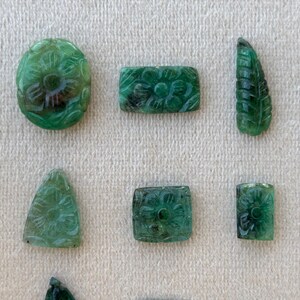 May include: Various carved green gemstones, including oval, rectangular, and leaf-shaped pieces. Each gemstone is intricately decorated with floral patterns, demonstrating detailed craftsmanship. The gemstones are arranged on a neutral, textured surface.
