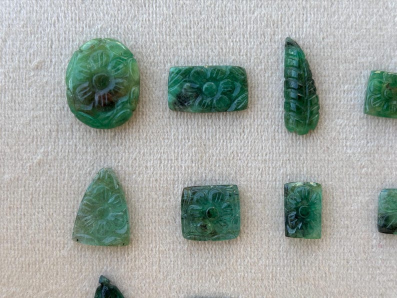 May include: Various carved green gemstones, including oval, rectangular, and leaf-shaped pieces. Each gemstone is intricately decorated with floral patterns, demonstrating detailed craftsmanship. The gemstones are arranged on a neutral, textured surface.
