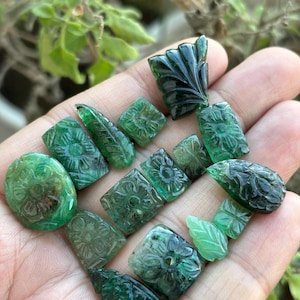 May include: A selection of carved emerald gemstones in diverse shapes, such as ovals, squares, and leaf designs. The gemstones are a deep green hue and feature detailed floral and leaf carvings. The gemstones are held in a hand.