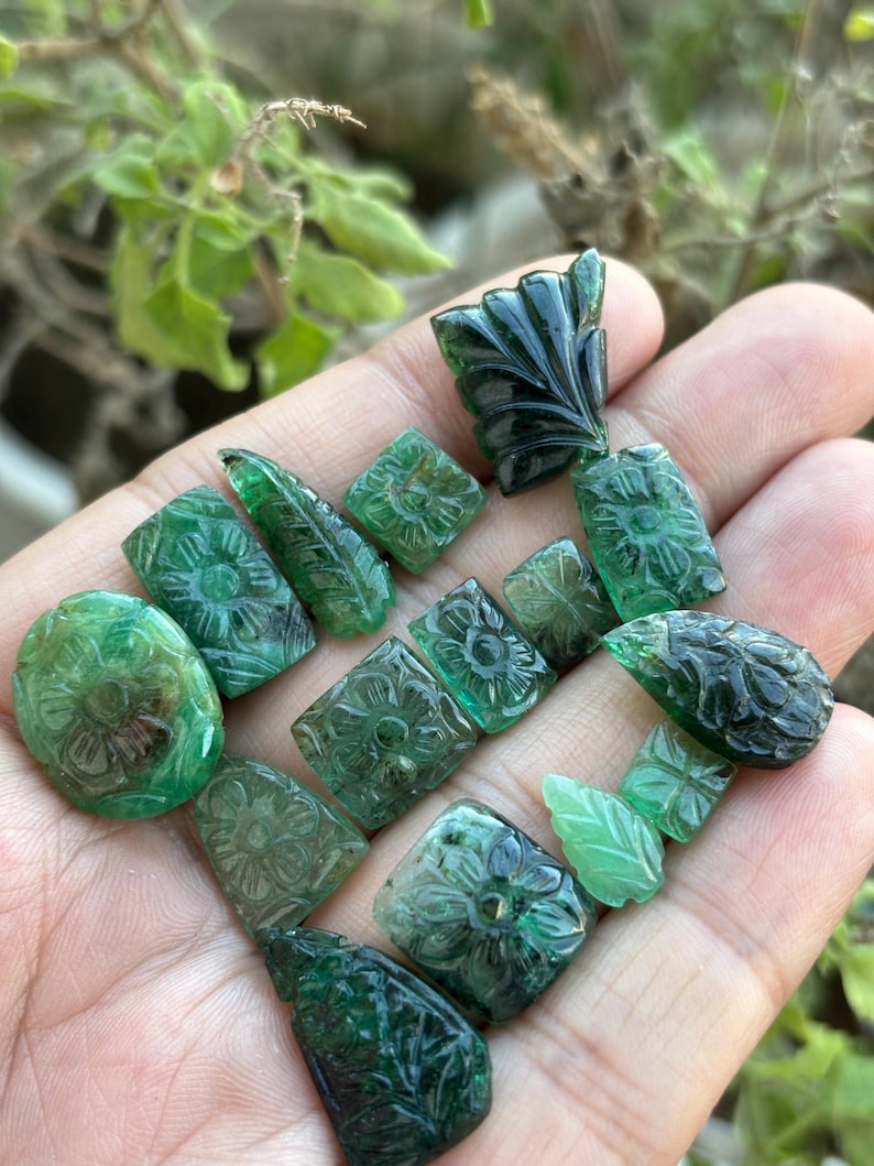 May include: A selection of carved emerald gemstones in diverse shapes, such as ovals, squares, and leaf designs. The gemstones are a deep green hue and feature detailed floral and leaf carvings. The gemstones are held in a hand.