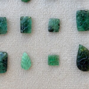May include: A collection of carved emerald gemstones in different shapes, including squares, rectangles, and a teardrop. The emeralds display floral and leaf patterns, with a variety of green shades.