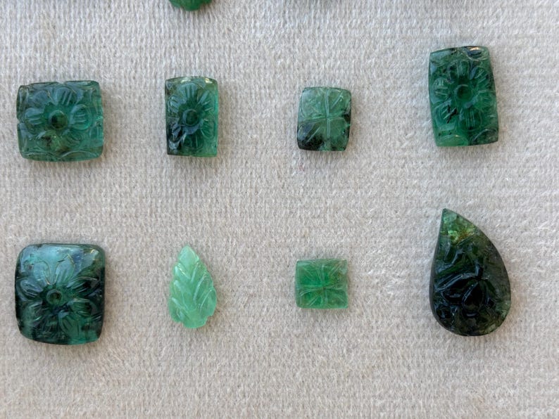 May include: A collection of carved emerald gemstones in different shapes, including squares, rectangles, and a teardrop. The emeralds display floral and leaf patterns, with a variety of green shades.