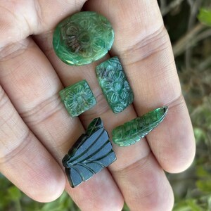 May include: A selection of carved green gemstones, including oval, rectangular, and square shapes, plus a leaf-shaped piece. The gemstones are held in an open hand, displaying their detailed floral and leaf patterns.