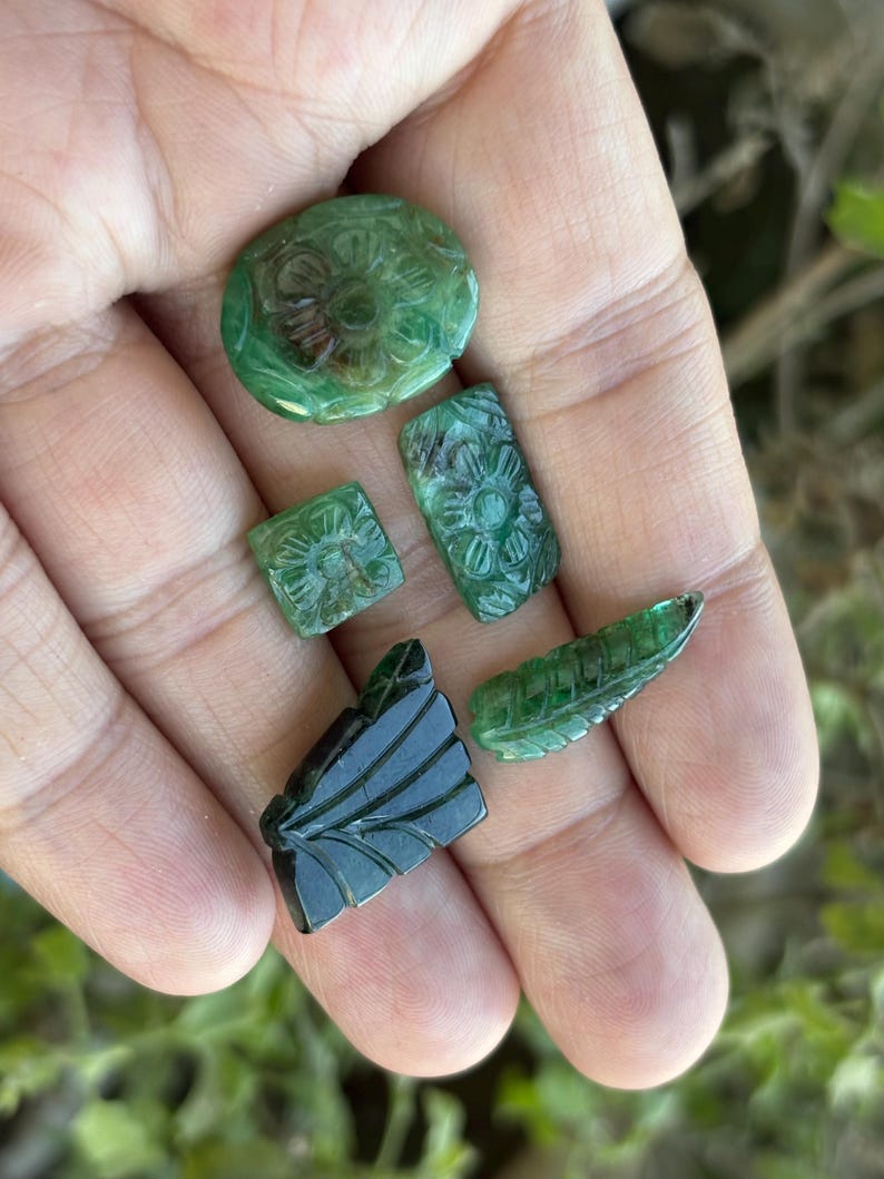May include: A selection of carved green gemstones, including oval, rectangular, and square shapes, plus a leaf-shaped piece. The gemstones are held in an open hand, displaying their detailed floral and leaf patterns.