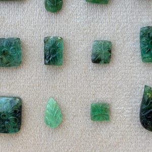 May include: A selection of carved emerald gemstones in diverse forms, such as squares, rectangles, and a leaf. The emeralds display floral and geometric patterns, exhibiting a spectrum of green shades. The gemstones are presented on a light-toned surface.