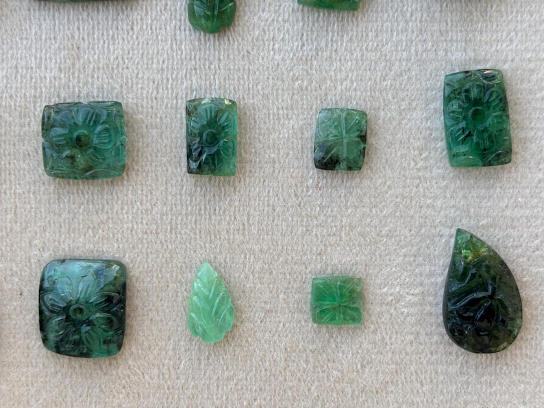 May include: A selection of carved emerald gemstones in diverse forms, such as squares, rectangles, and a leaf. The emeralds display floral and geometric patterns, exhibiting a spectrum of green shades. The gemstones are presented on a light-toned surface.