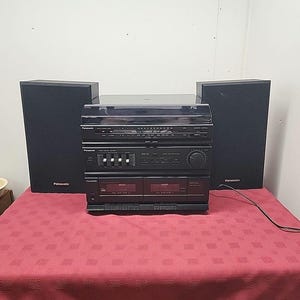 Puede incluir: Un equipo estéreo Panasonic vintage con dos altavoces negros y una unidad central con radio, reproductores de casetes y un tocadiscos. El sistema está sobre un mantel rojo estampado.