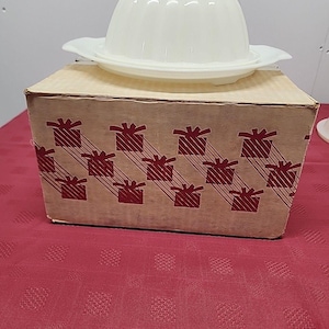 Puede incluir: Un molde de gelatina vintage de dos piezas, de color blanquecino, con tapa, sobre un plato a juego. El molde está encima de una caja de cartón con un patrón repetido de cajas de regalo. La caja está sobre un mantel rojo.