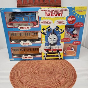 May include: A Thomas the Tank Engine & Friends battery operated railway toy set in its original blue cardboard packaging. The box displays a blue Thomas engine, brown Annie and Clarabel passenger coaches, and text detailing the set contents.