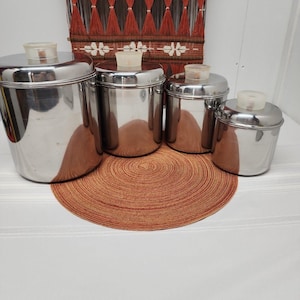 Könnte beinhalten: Ein Set aus vier zylindrischen Vorratsdosen aus Edelstahl mit polierten, reflektierenden Oberflächen und passenden Deckeln. Jeder Deckel ist mit einem kleinen, durchscheinenden, cremeweißen Knaufgriff in der Mitte versehen.