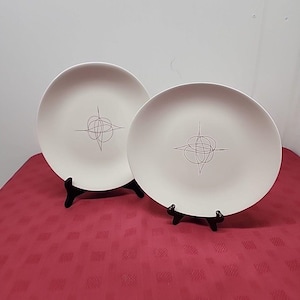 May include: Two off-white ceramic plates with a gray abstract design. The plates are displayed on black stands against a red tablecloth. The design on each plate is a gray, stylized starburst.