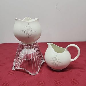 May include: A white ceramic creamer and sugar bowl set. The creamer has a handle and spout, while the sugar bowl sits atop a clear glass pedestal. Both pieces feature a black geometric design.