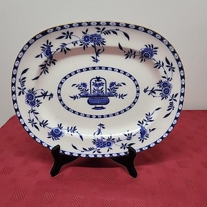 May include: Oval serving platter with a white base and blue floral design. The platter features a central oval motif with a floral basket and is bordered by a repeating floral pattern. The rim is accented with a gold trim.